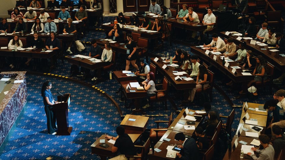 Student senators seated at desks in a full replica of the United States Senate chamber, one speaking from the floor.